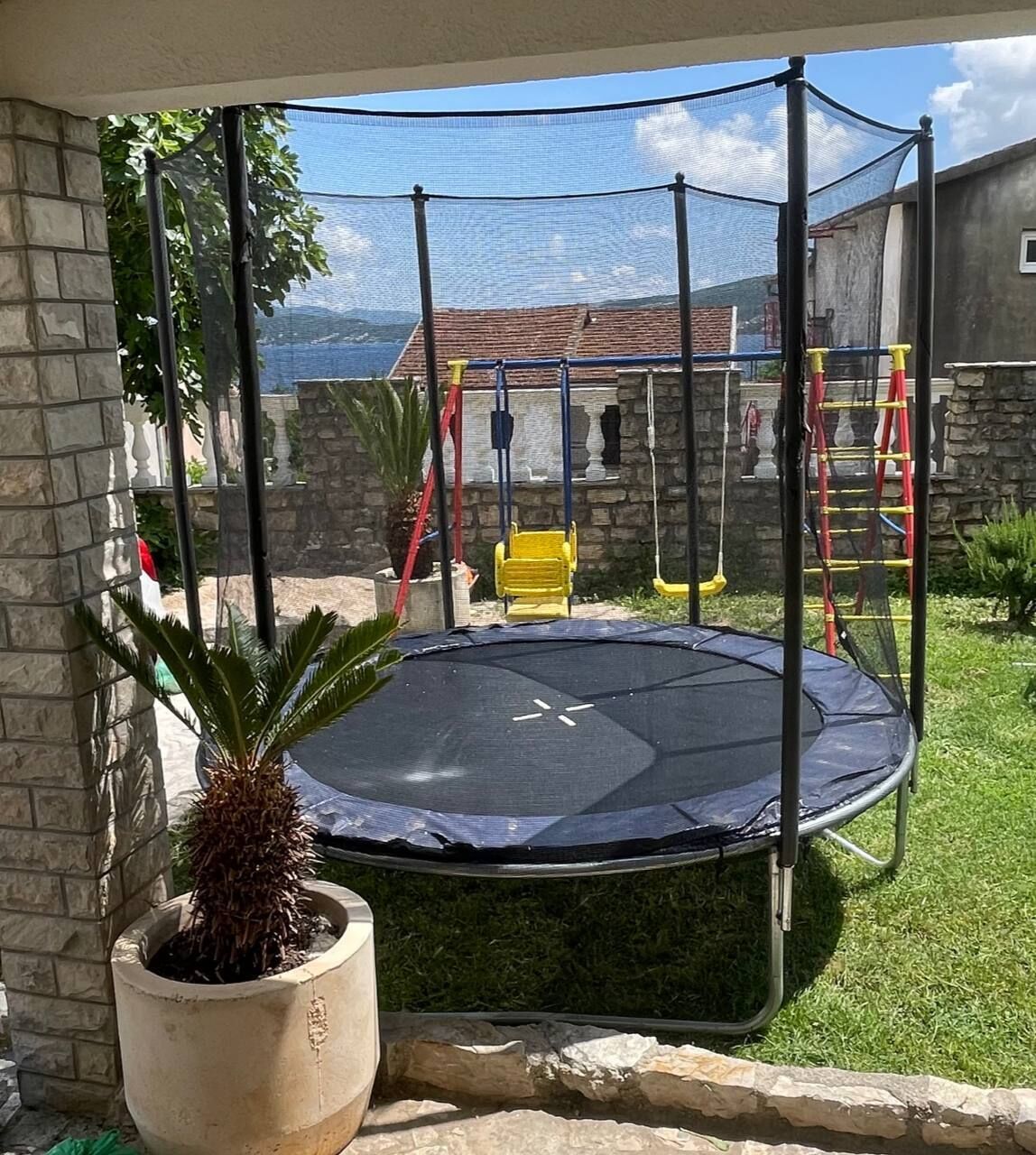 Un trampolín en un patio trasero con un muro de piedra, una planta en maceta y un conjunto de columpios.