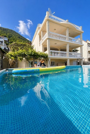 Una piscina amb una balsa inflable groga, un edifici blanc amb balcons i un turó verd al fons.