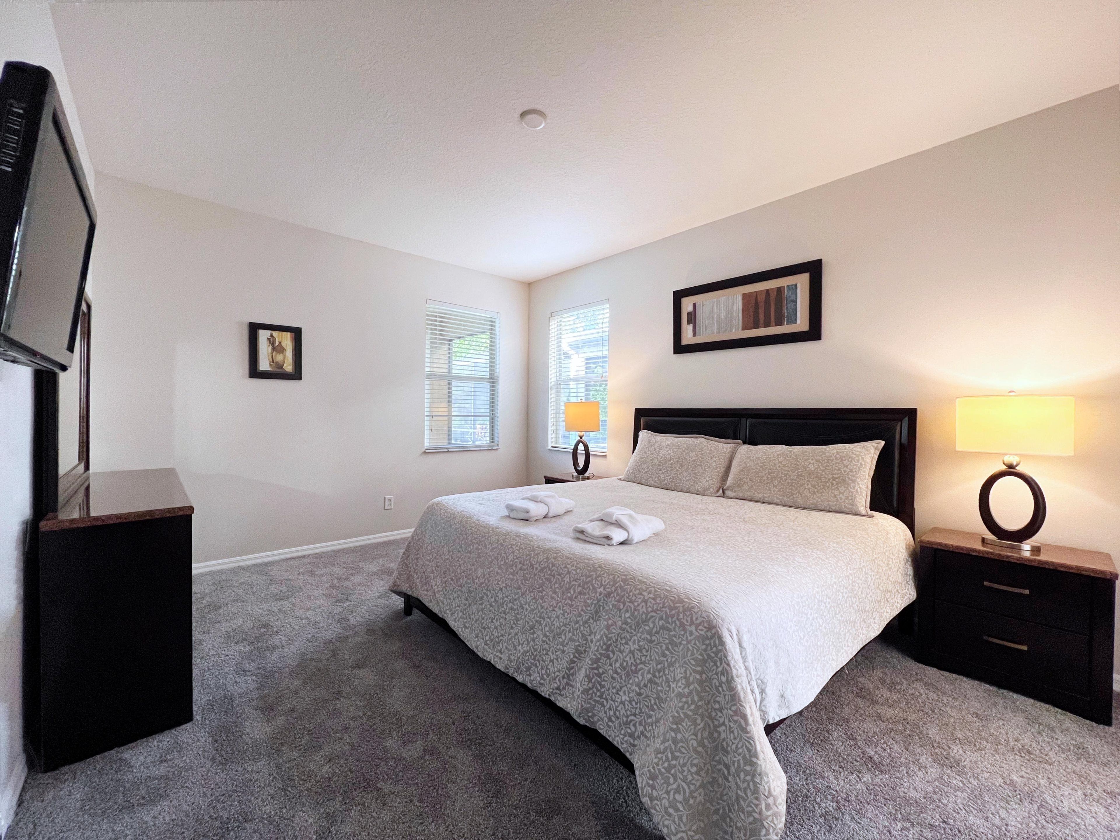 A bedroom with a bed, two bedside tables, a TV, and a window with blinds.