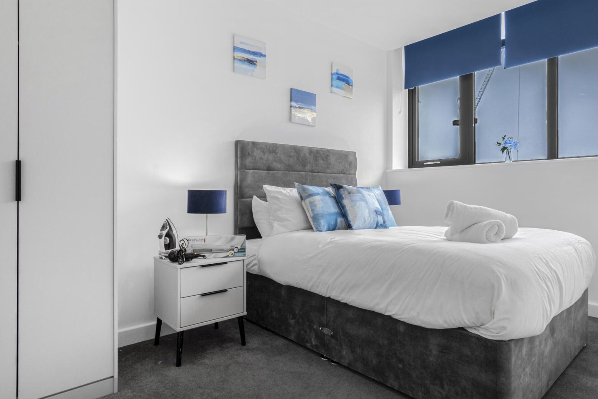 Un dormitorio moderno con una cama tapizada de gris, ropa de cama blanca y una mesita de noche con lámpara. Hay tres cuadros colgados en la pared sobre la cama.