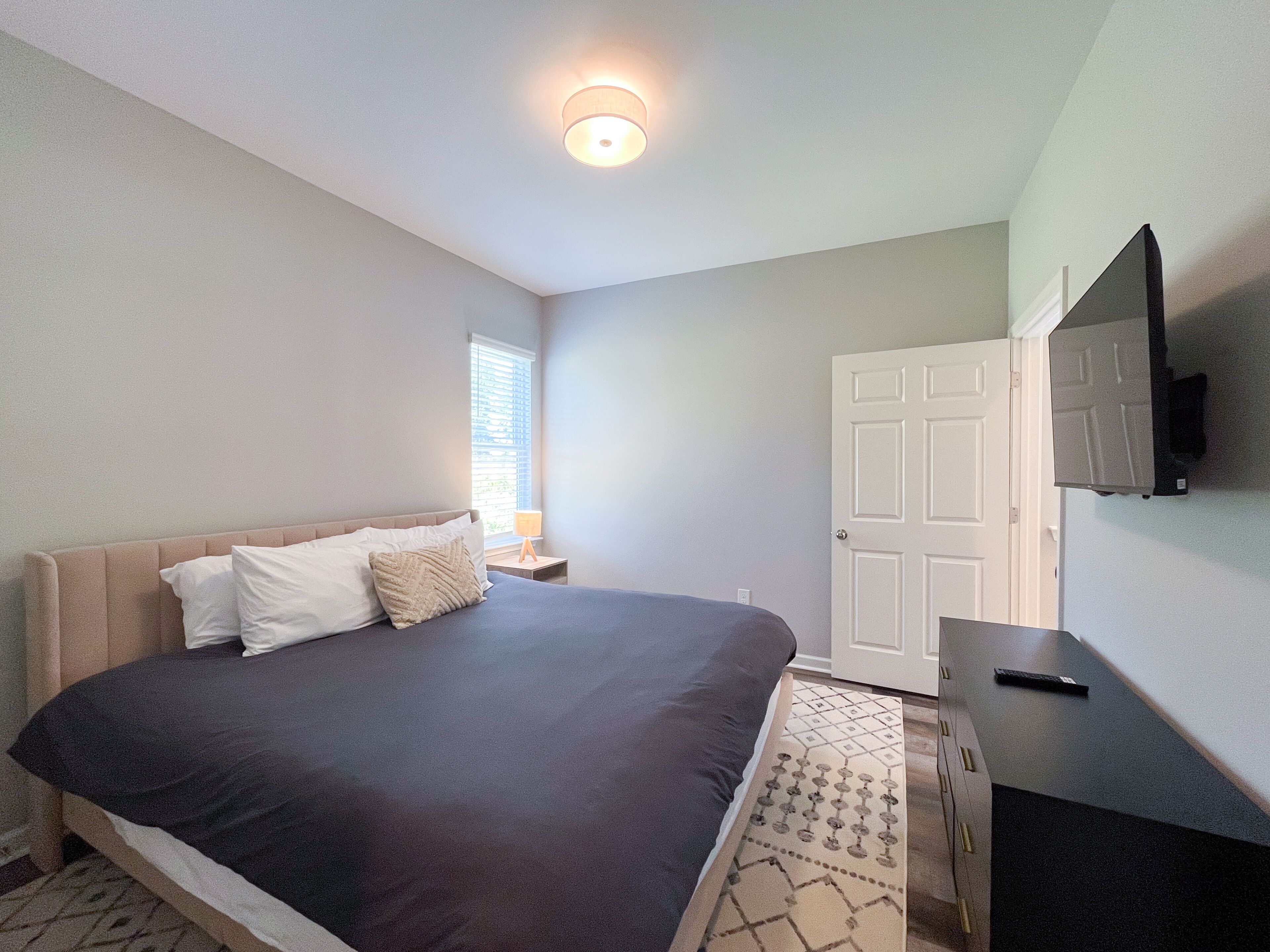 A bedroom with a bed, a bedside table with a lamp, a TV mounted on the wall, and a door.