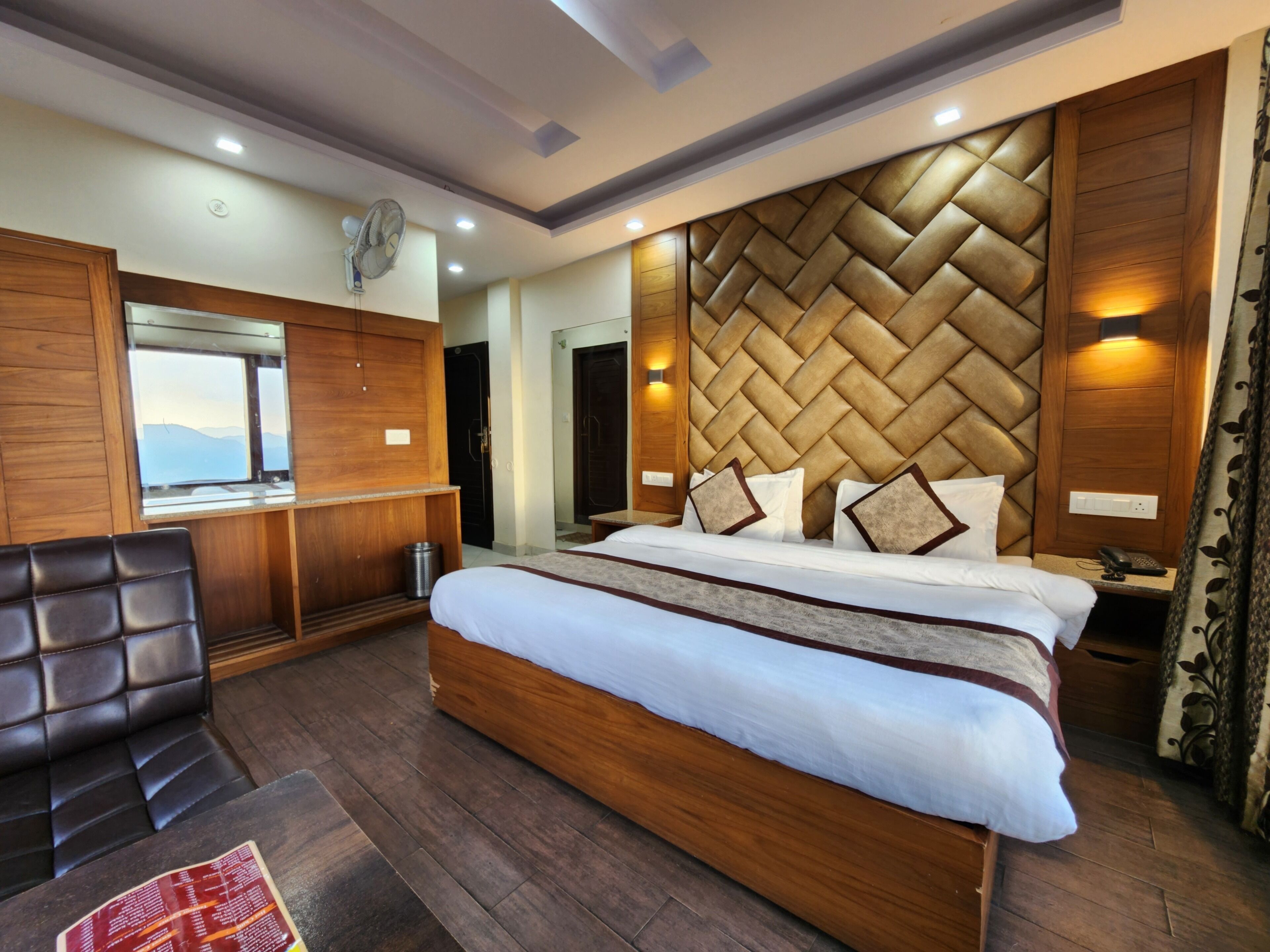 A modern hotel room with a large bed, wooden headboard, a seating area with a chair, and a view of the mountains through a window.