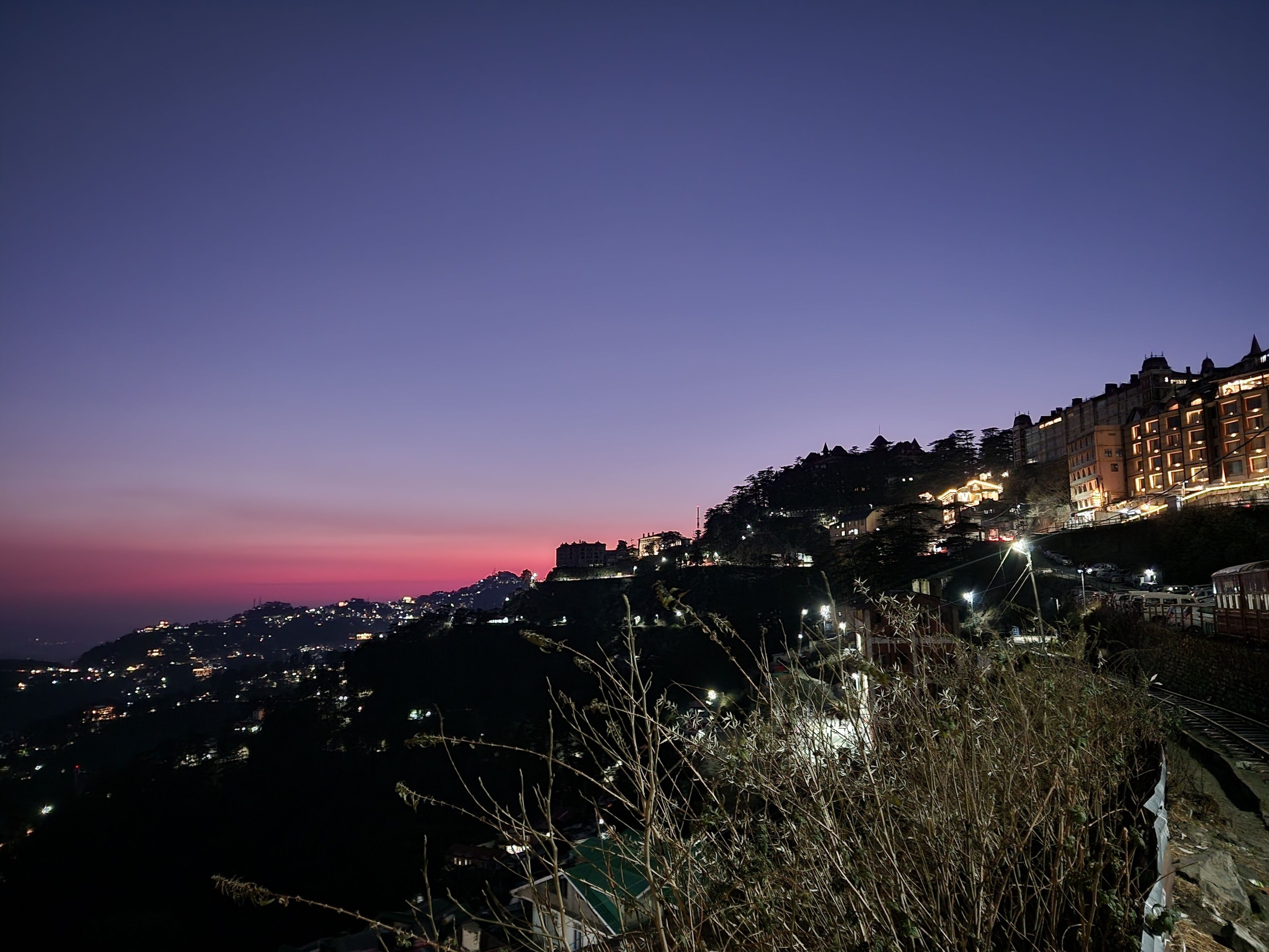 A nighttime view of a hillside town with illuminated buildings and a train track.