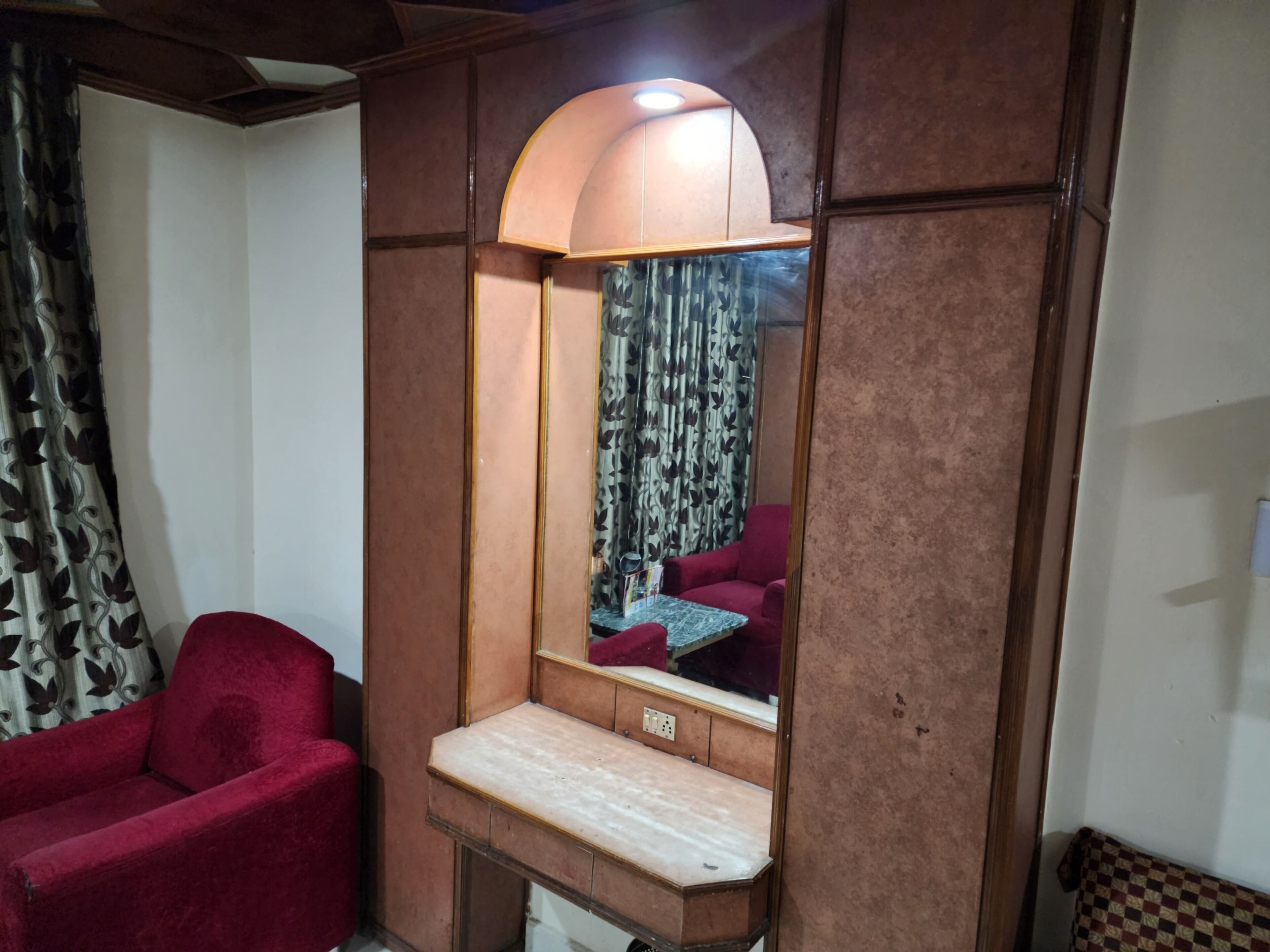 A room with a red armchair, a wooden cabinet with a mirror, and patterned curtains.