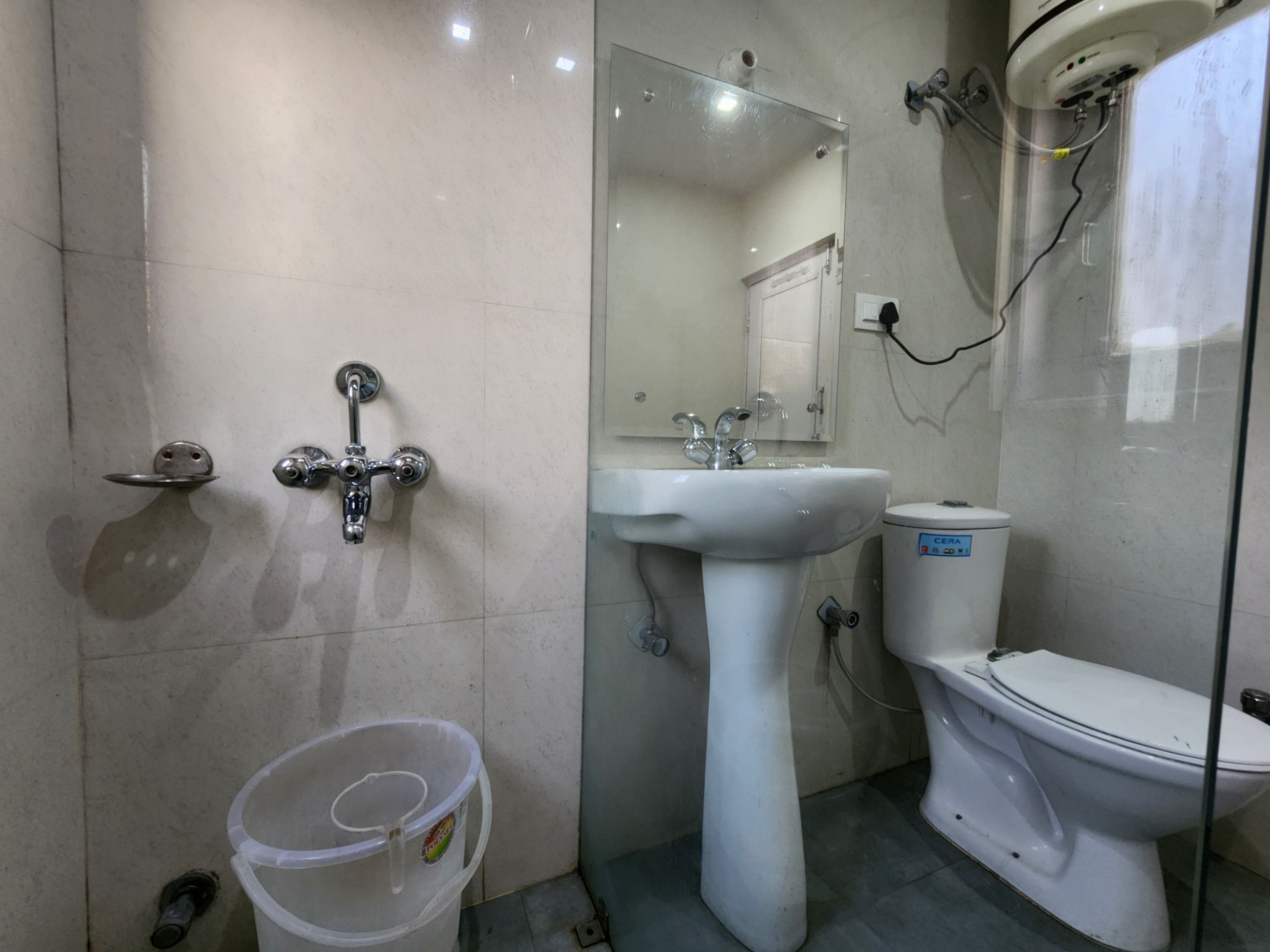 Ein Badezimmer mit weißem Waschbecken, WC und Dusche. Auf dem Boden steht ein Plastikeimer.