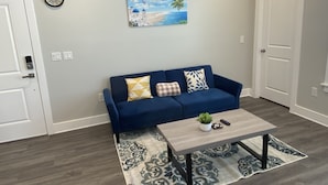 Een woonkamer met een blauwe bank, een salontafel, een schilderij van een strandgezicht en een klok aan de muur.