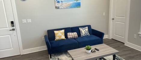 A living room with a blue sofa, a coffee table, a painting of a beach scene, and a clock on the wall.