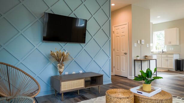 A modern living room with a mounted TV, wicker furniture, and a kitchen in the background.