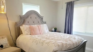 A bedroom with a tufted headboard, a bed with a floral bedspread, a bench, a nightstand with a plant, and a window with curtains. - Adorable Downtown Mesa Cottage (Mesa)
