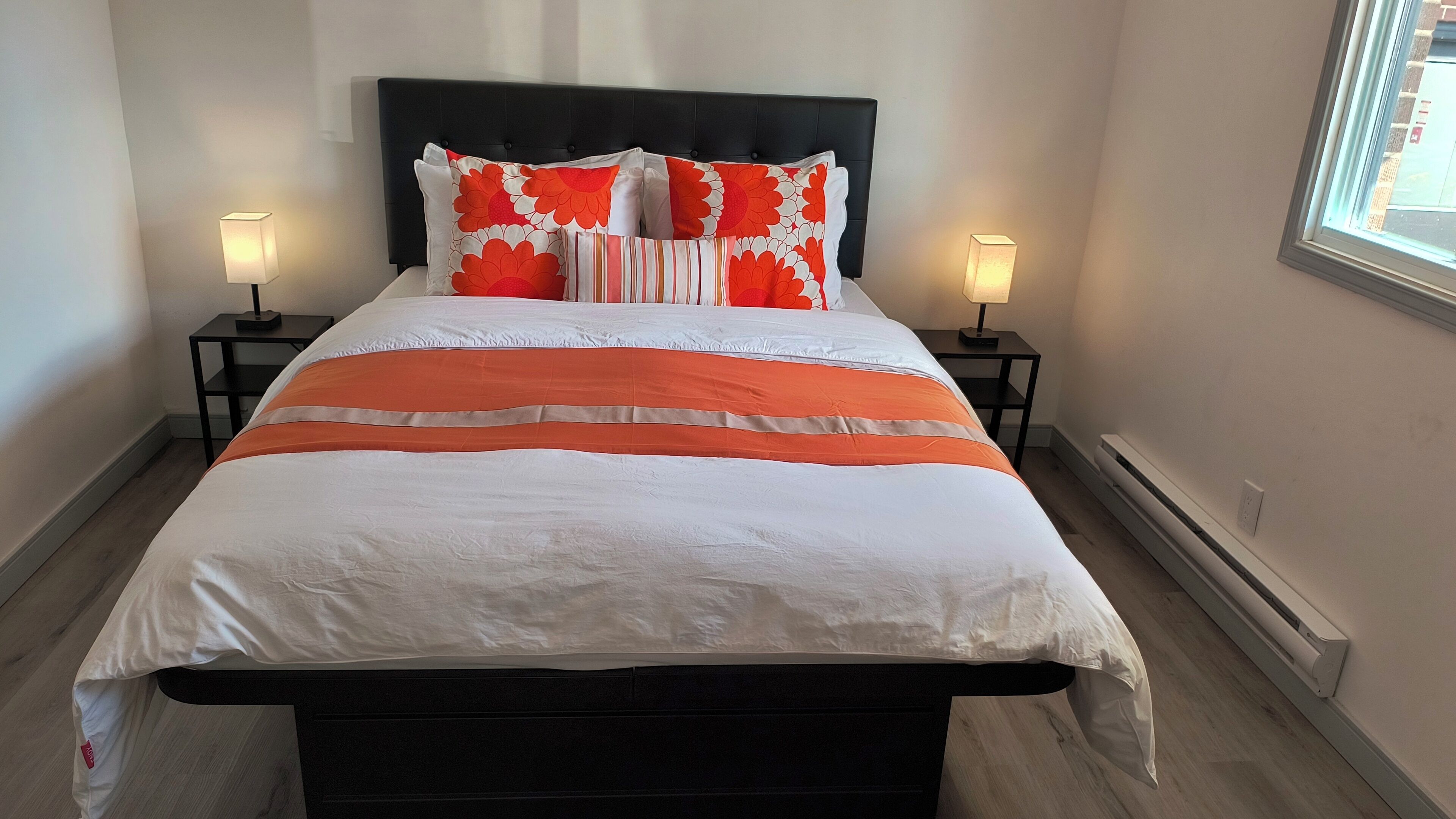 A bed with a white and orange bedspread, two nightstands with lamps, and a window with a view of the street.