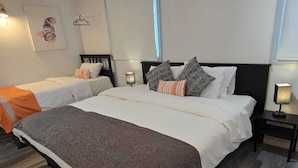 A hotel room with two beds, a large bed with a brown blanket and a smaller bed with an orange blanket. There are two bedside tables with lamps, a wall-mounted light, and a painting on the wall.