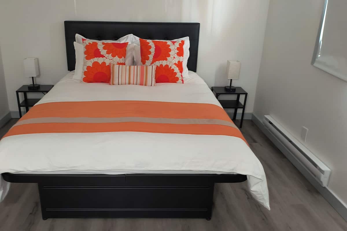 A modern bedroom with a black headboard, a large bed with white and orange bedding, two bedside tables with lamps, and a wooden floor.