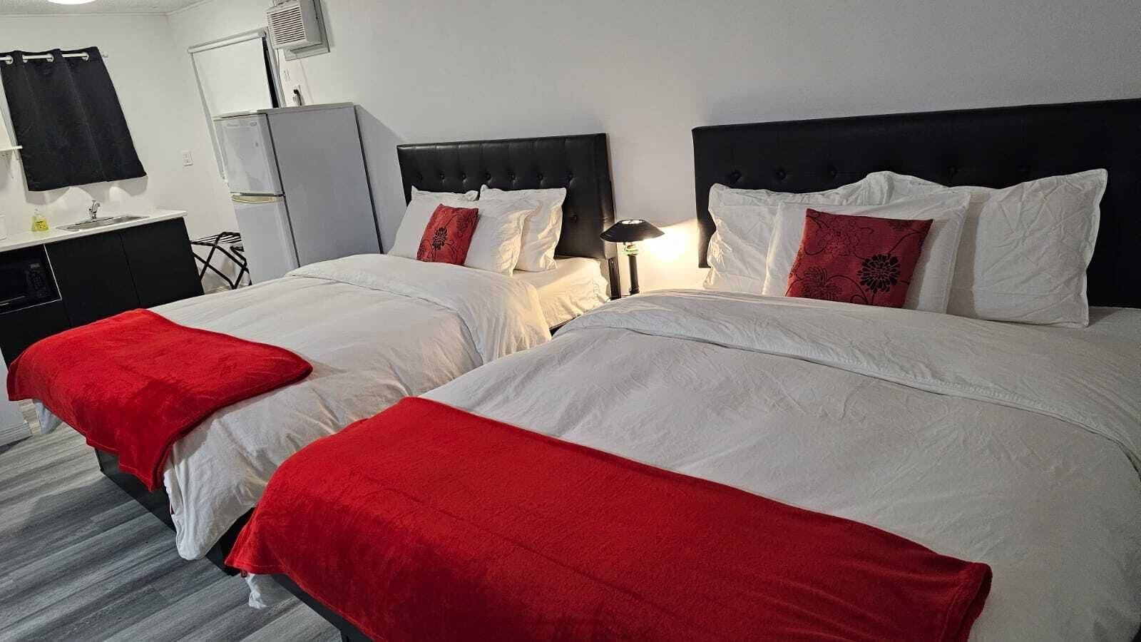 A hotel room with two beds, each with white bedding and red accents. There is a small desk with a lamp, a wardrobe, and a refrigerator.