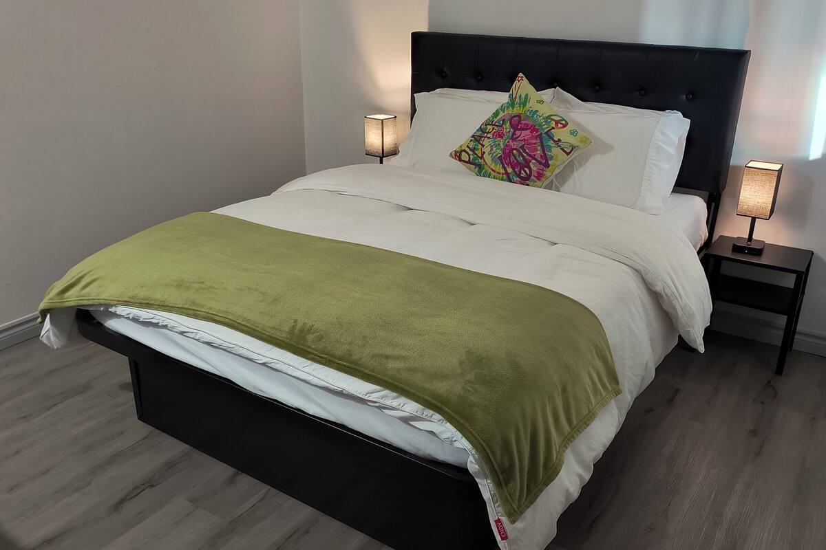 A neatly made bed with a green throw blanket, white pillows, and a colorful pillow. There are two bedside tables with lamps, a dark headboard, and a light-colored wall.