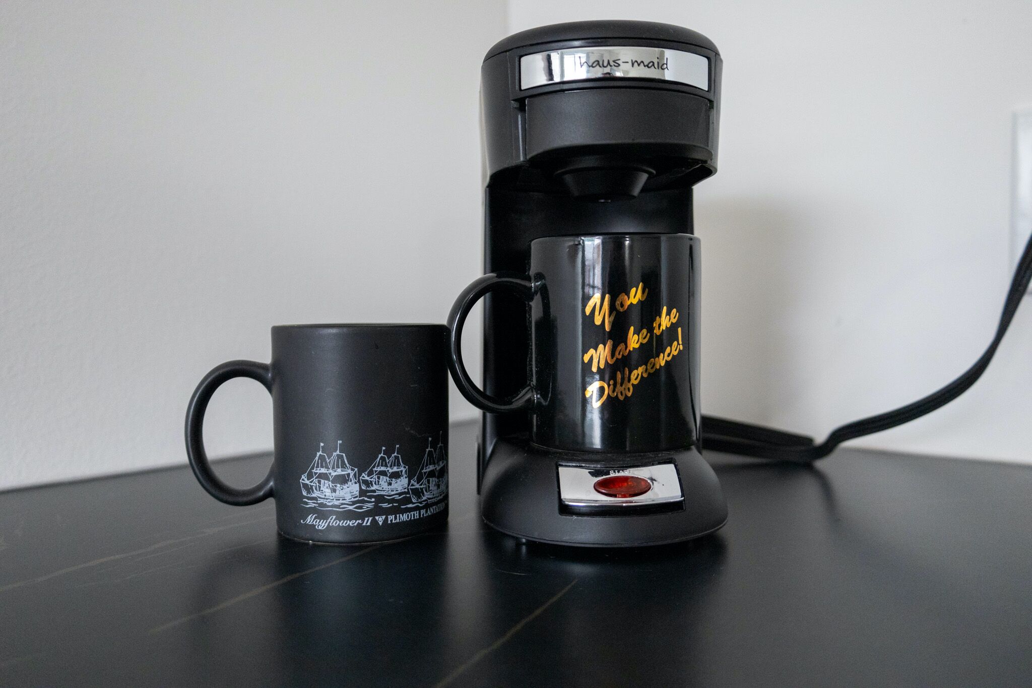 Uma cafeteira preta e uma caneca preta com o desenho de um barco e texto sobre uma mesa.