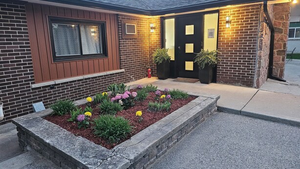 A building with a brick facade, a black door with a window, and a flower bed with various flowers.
