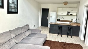 A modern living room with a grey sofa, a brown rug, and a kitchen area with a white refrigerator, black stools, and a coffee maker. - Saint Sebastian Flat 603Duplex no Centro (Jaragua do Sul)