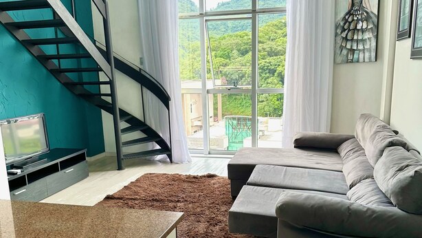 A modern living room with a staircase, a sofa, a coffee table, and a view of greenery through large windows. - Saint Sebastian Flat 603Duplex no Centro (Jaragua do Sul)