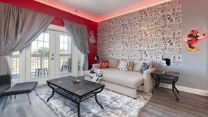 A living room with a red accent wall, a large window with curtains, a sofa, a coffee table, and a wall decorated with cartoon character prints. - Disney themed 3br condo (Kissimmee)