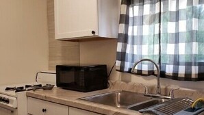 A compact kitchen with white cabinets, a black microwave, a stainless steel sink, and a window with checkered curtains. - Private Studio whole unit (Rockford)
