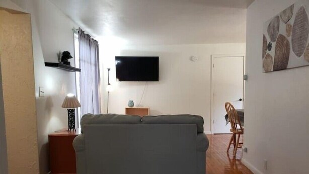 A compact living space with a sofa, a flat-screen TV mounted on the wall, a small table with a lamp, and a chair. - Private Studio whole unit (Rockford)