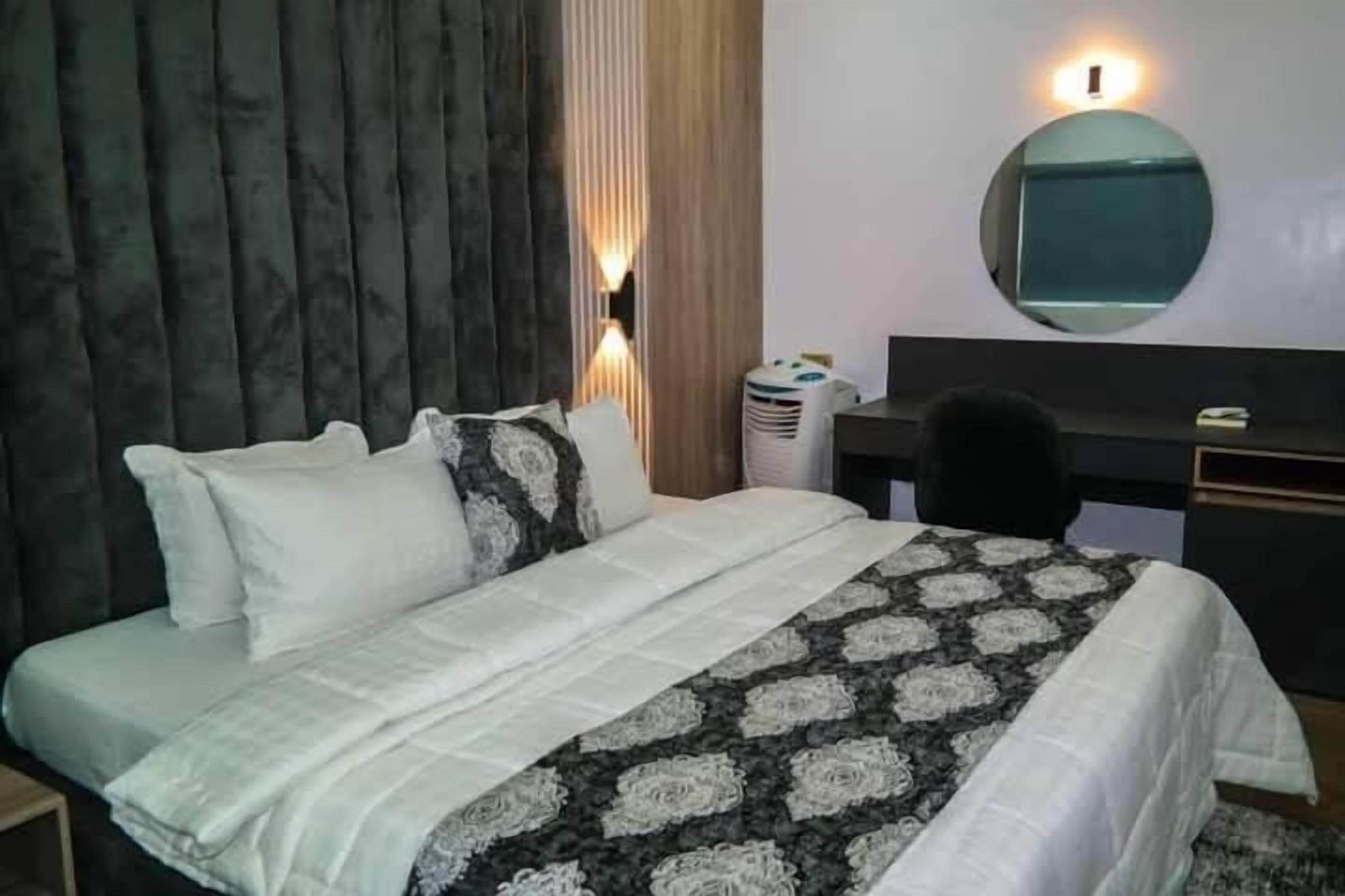 A hotel room with a bed, a desk, a chair, a mirror, and a wall-mounted light.