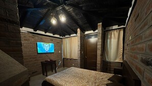 A bedroom with a brick wall, a wooden ceiling, a bed with a patterned bedspread, a television on the wall, and a door with curtains.