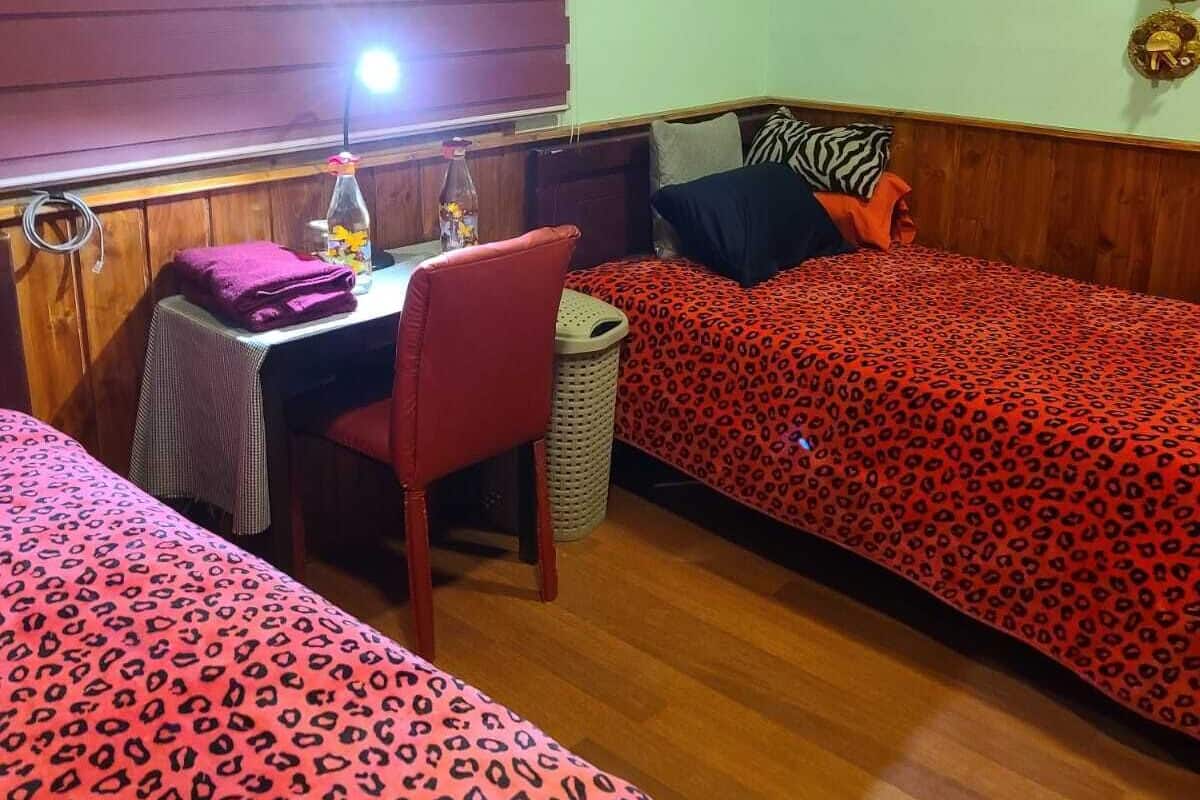 A room with two beds, a small table with a lamp, a chair, and a wall-mounted shelf with decorative items.