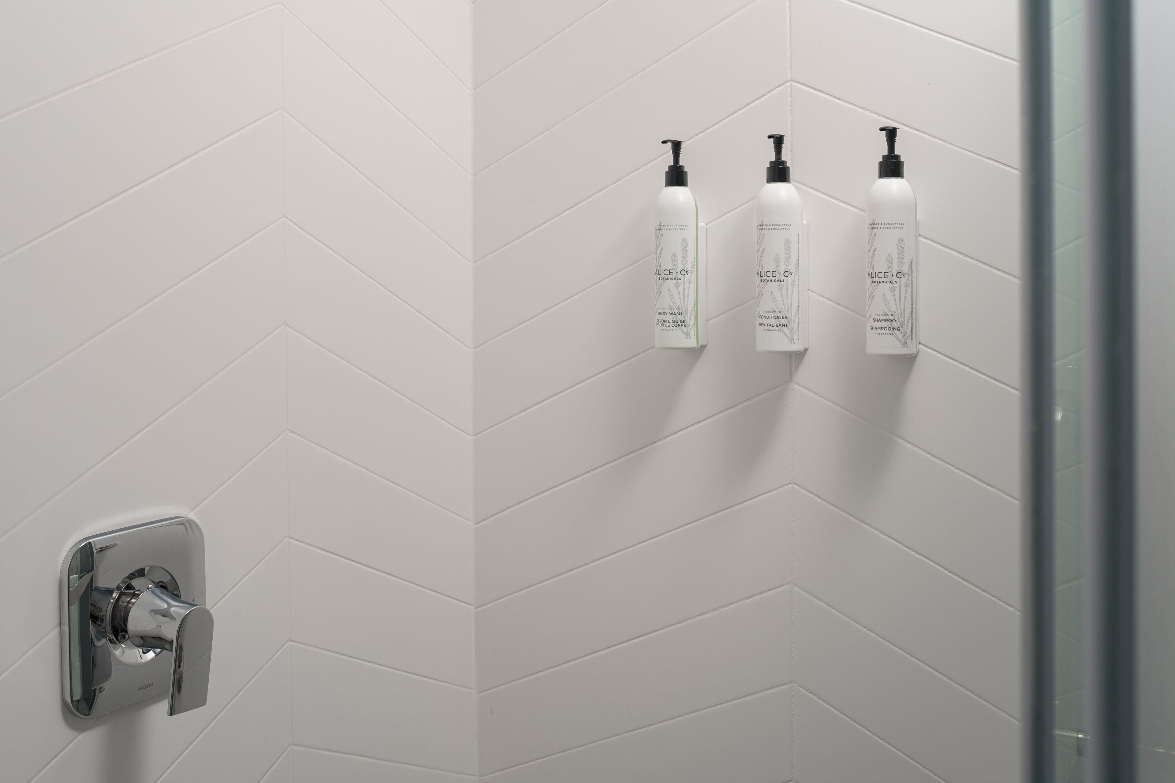 A bathroom with white herringbone tiles, a shower with a chrome faucet, and three bottles of Alice + Co body wash and shampoo on the wall.