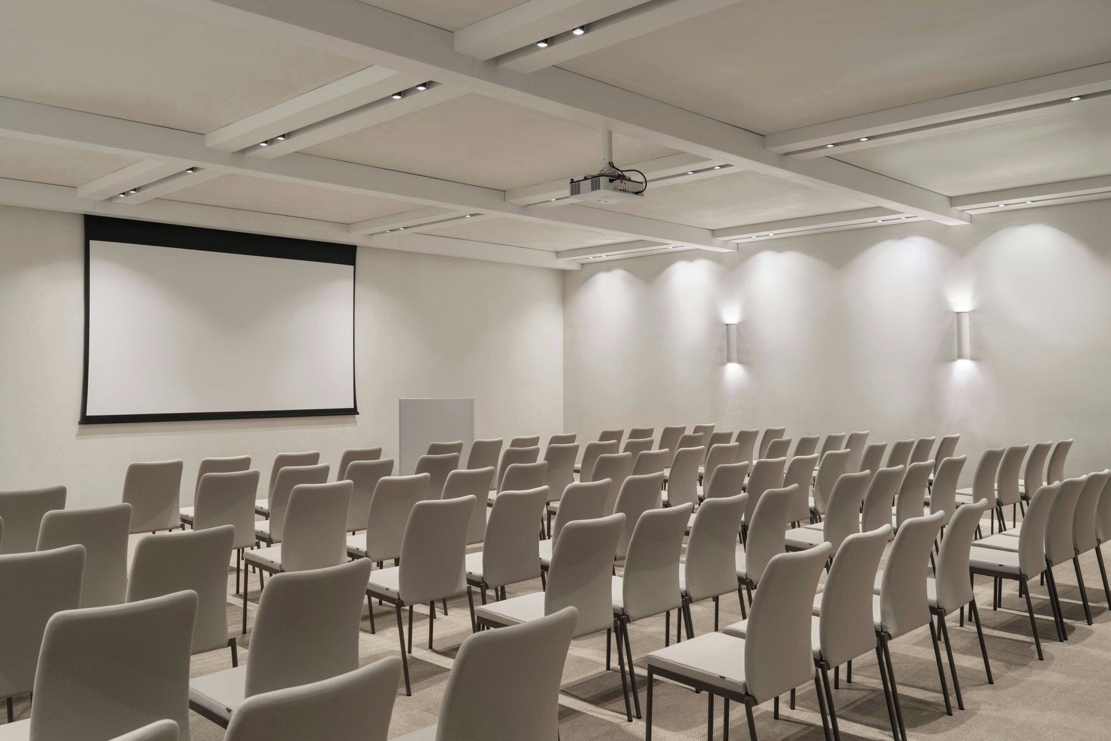 A conference room with rows of chairs and tables, a projector screen, and ceiling lights.