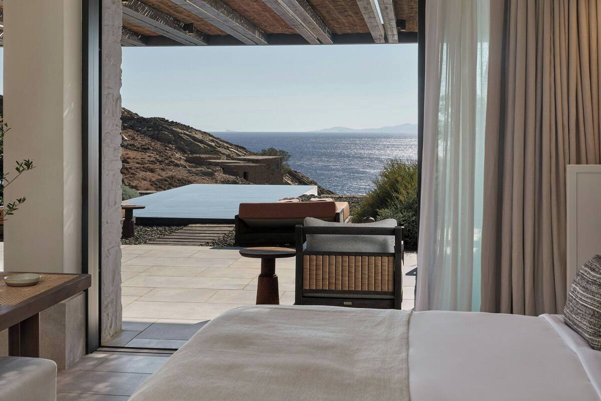 A bedroom with a large bed, a small table, and a chair. There is a view of the sea and a rocky landscape outside the window.