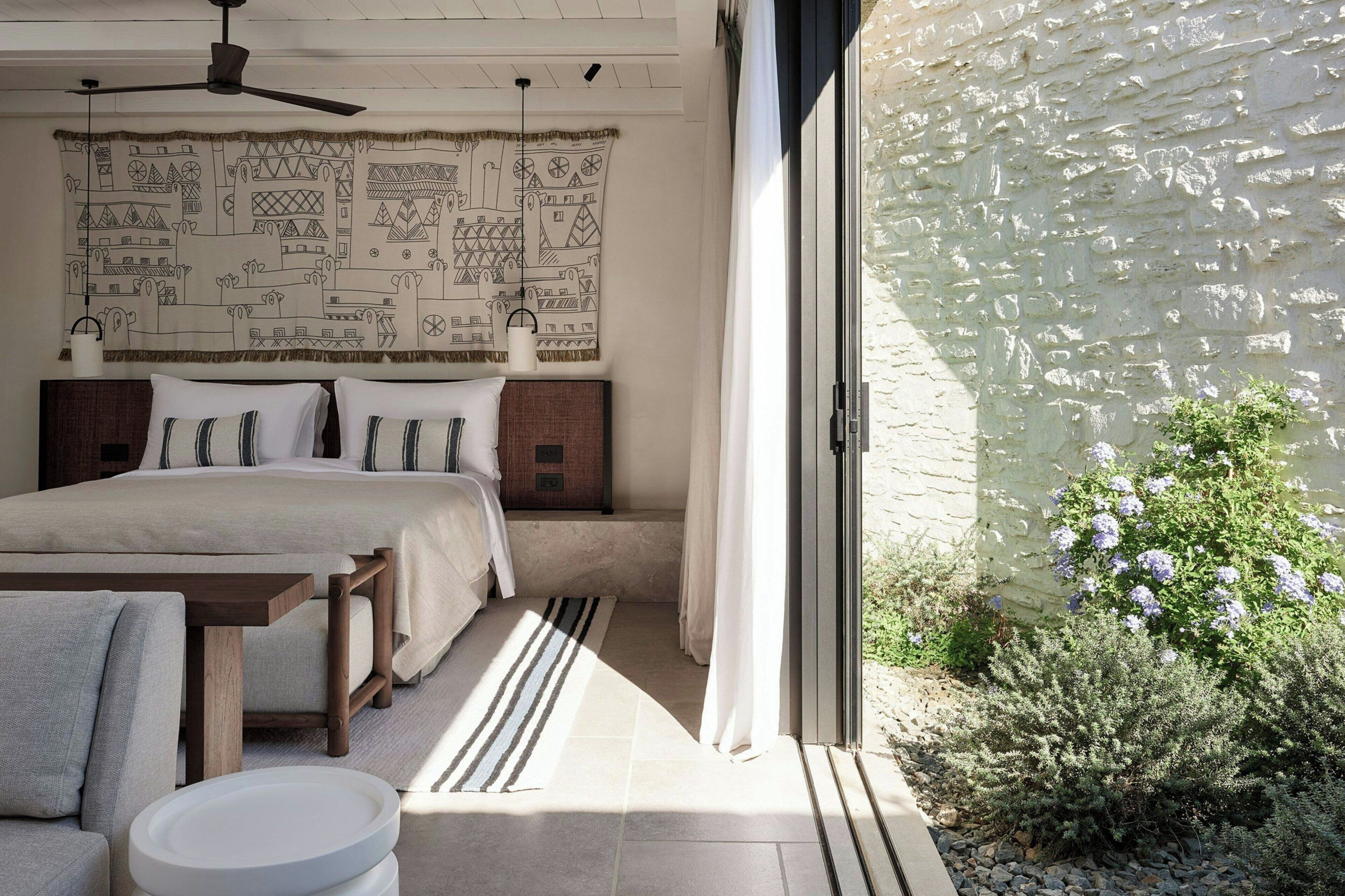 A bedroom with a bed, bedside tables, a ceiling fan, and a view of a garden through a glass door.