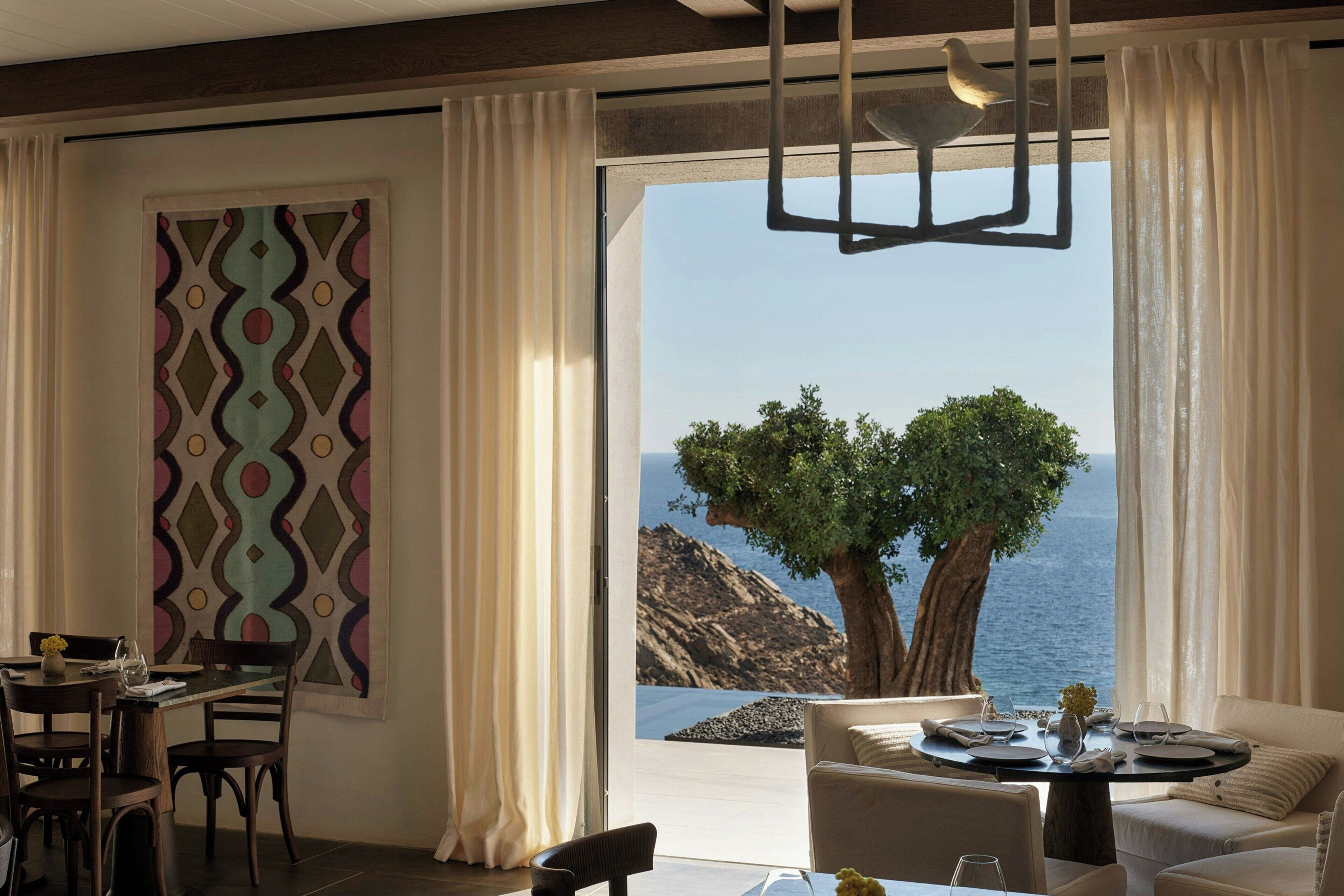 Un espace de restauration moderne offrant une vue sur la mer, agrémenté d’un grand arbre et d’une œuvre d’art géométrique murale.