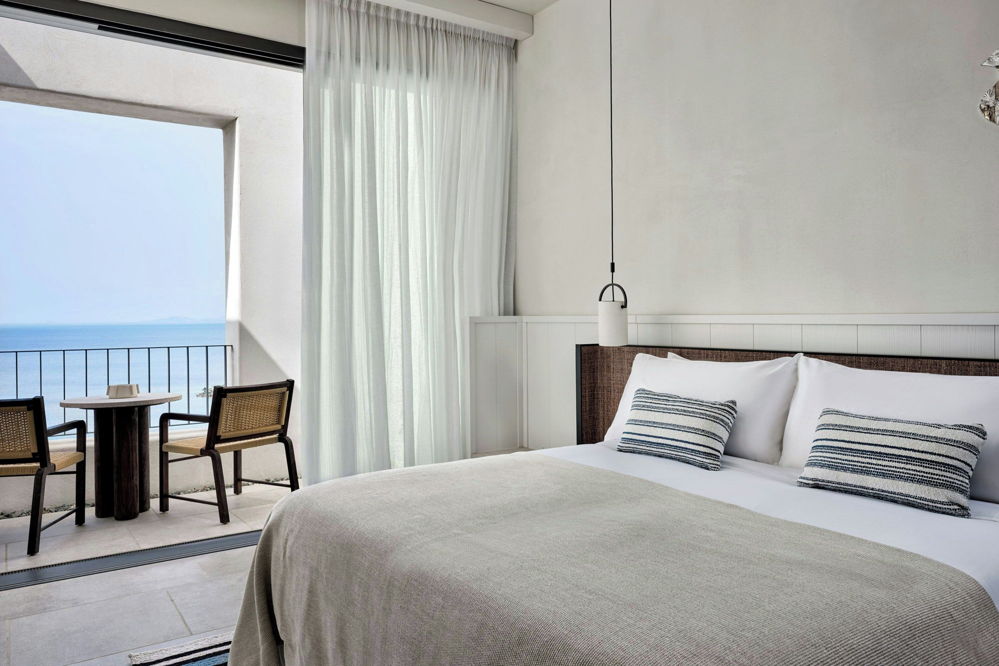 A hotel room with a large bed, two pillows, a view of the sea, a balcony with a table and chairs, and a hanging light fixture.