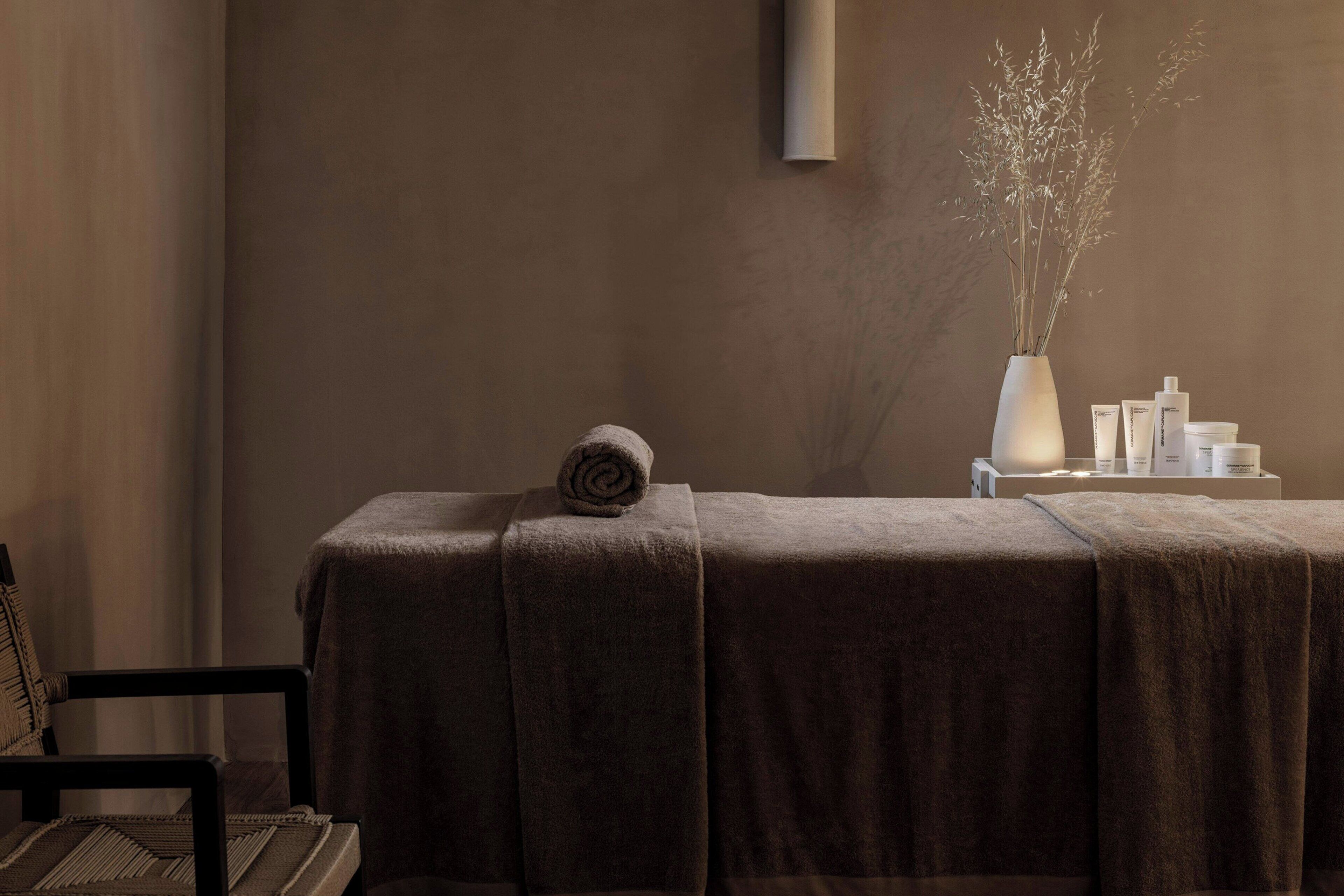 A spa area with a massage table covered in a brown towel, a rolled towel on the table, a vase with dried plants, and various spa products on a small table beside the table.