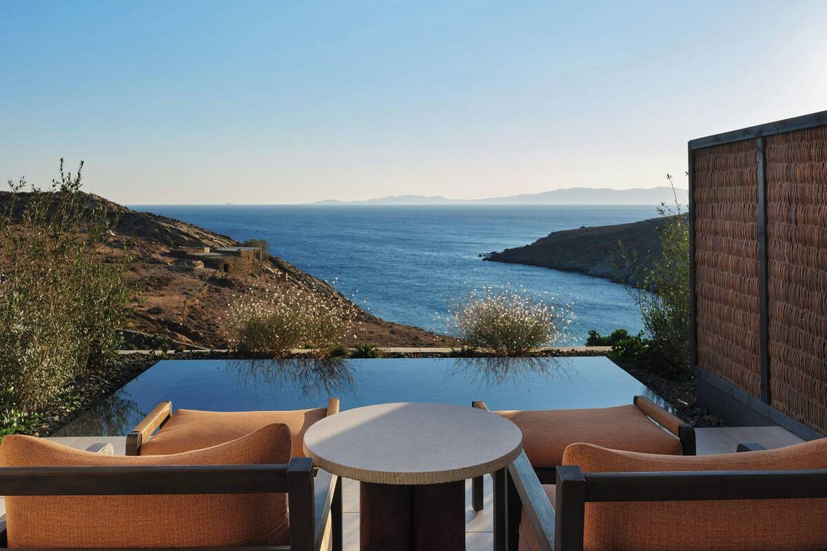 An infinity pool with a view of the ocean and hills. There are two chairs and a small table.