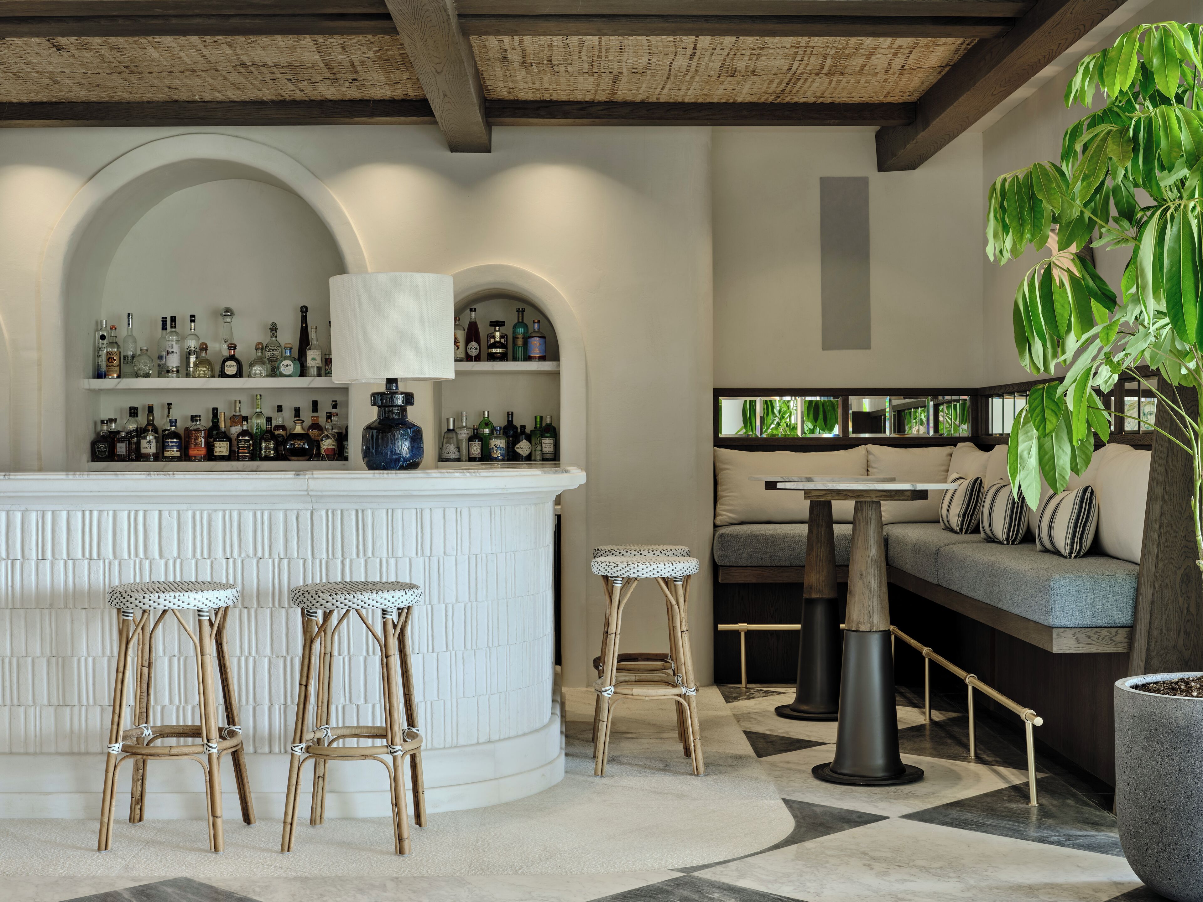 Una zona de bar con un mostrador blanco, estantes llenos de botellas de licores y dos taburetes.