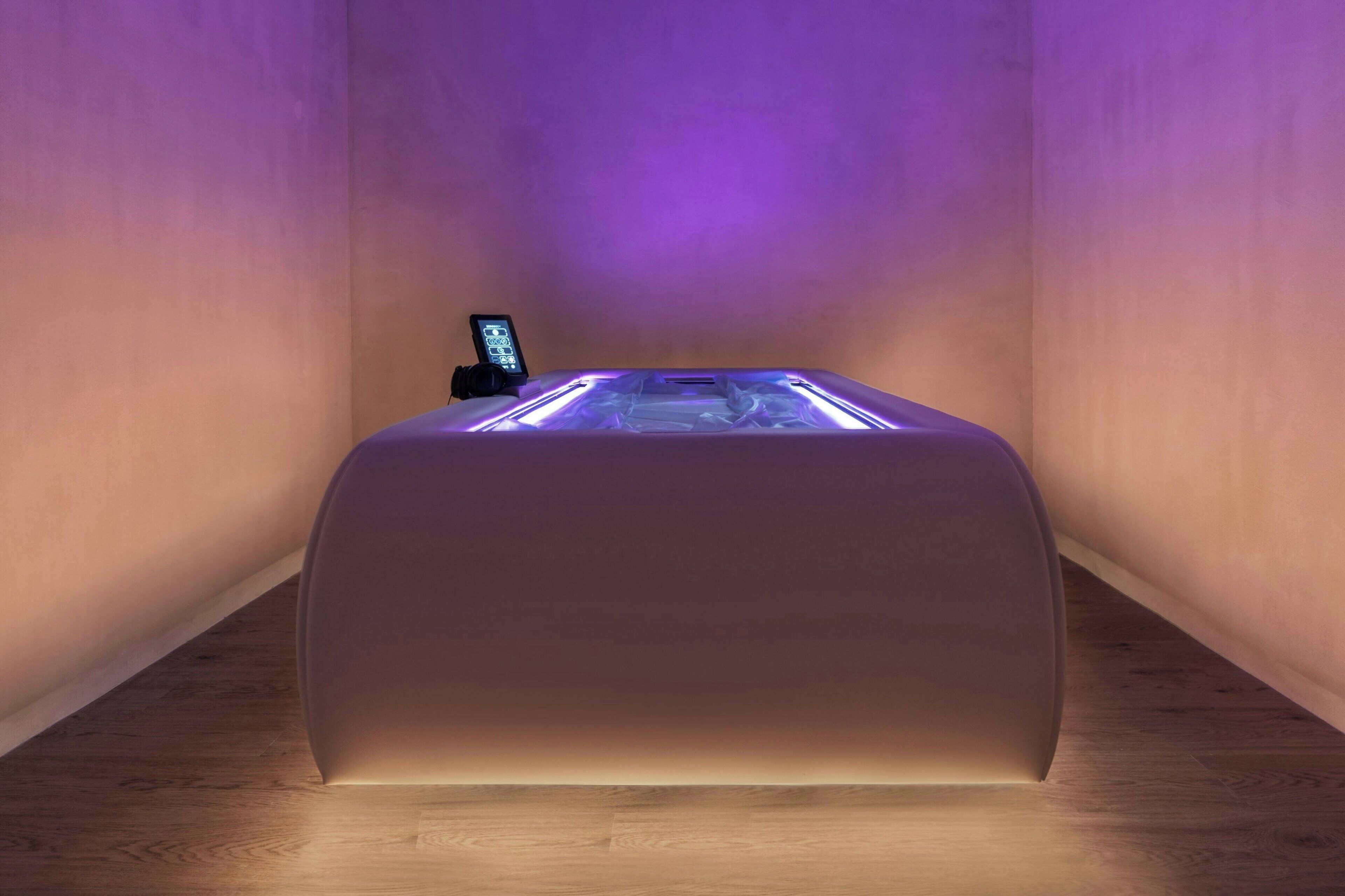A modern, minimalist room with a central, illuminated, glass-top table and a control panel on the left.