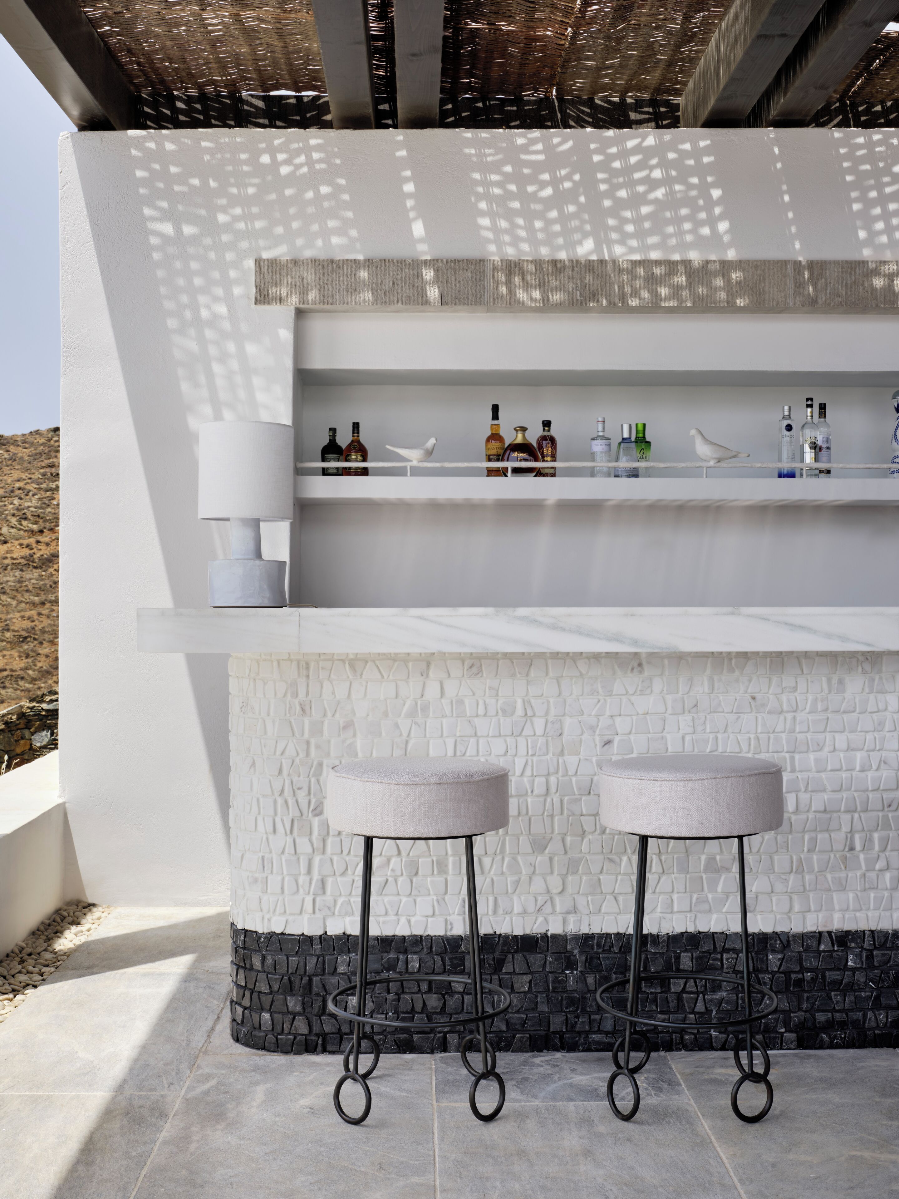 A modern outdoor bar area with two stools, a white textured wall, and a shelf with various bottles.