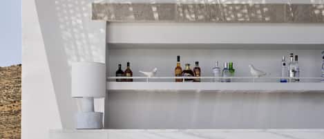 A modern outdoor bar area with two stools, a white textured wall, and a shelf with various bottles.