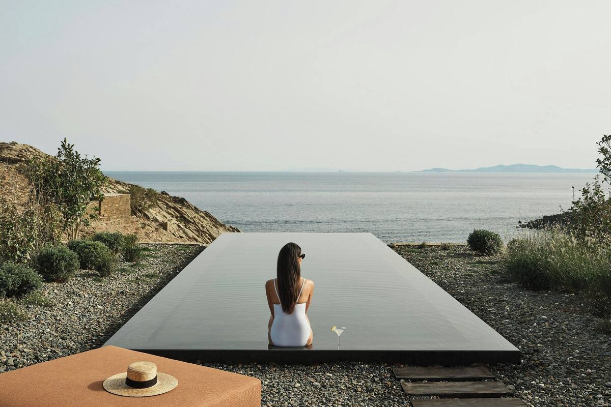 A woman sitting on a long, flat surface by the sea, with a hat on a table in the foreground.