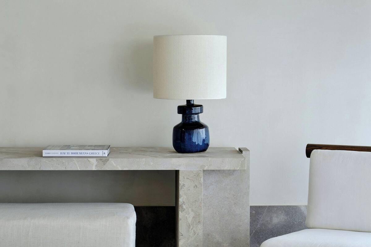 A modern room with a marble console table, a blue lamp, a book titled "HAT TE BOHEMIANS GREECE", and a white chair.