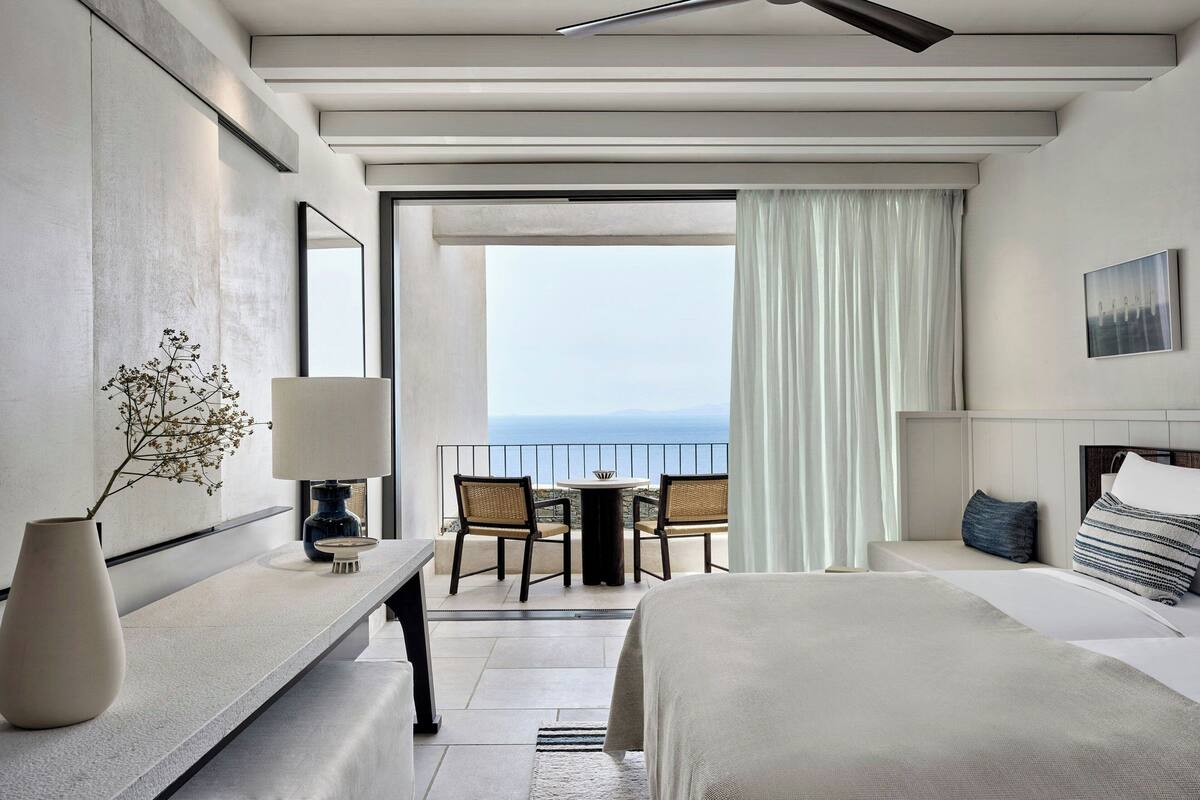 A modern hotel room with a balcony, a bed, a desk, and a view of the sea.