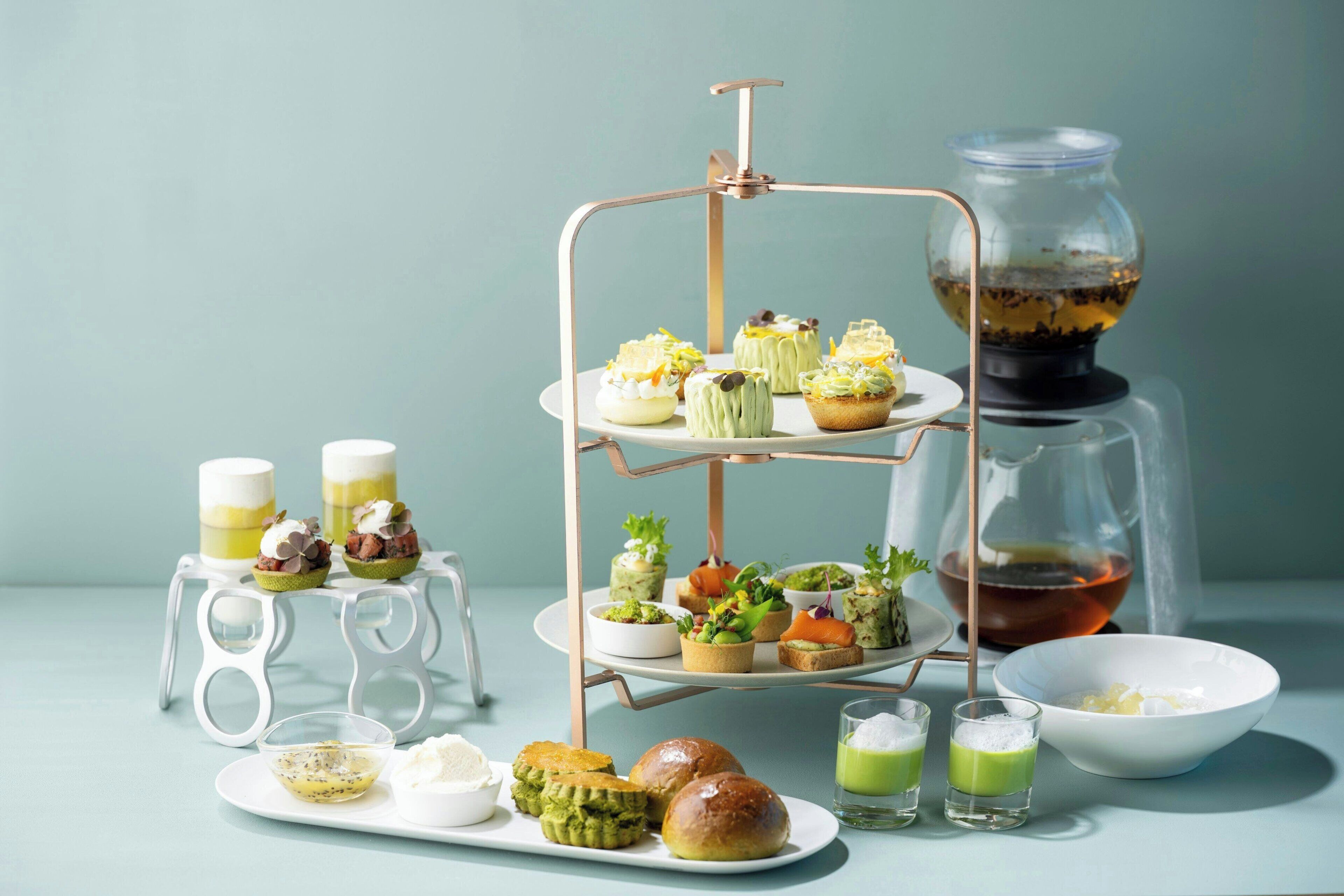 A tiered serving tray with various food items, a coffee machine, and a glass of tea on a light blue surface.