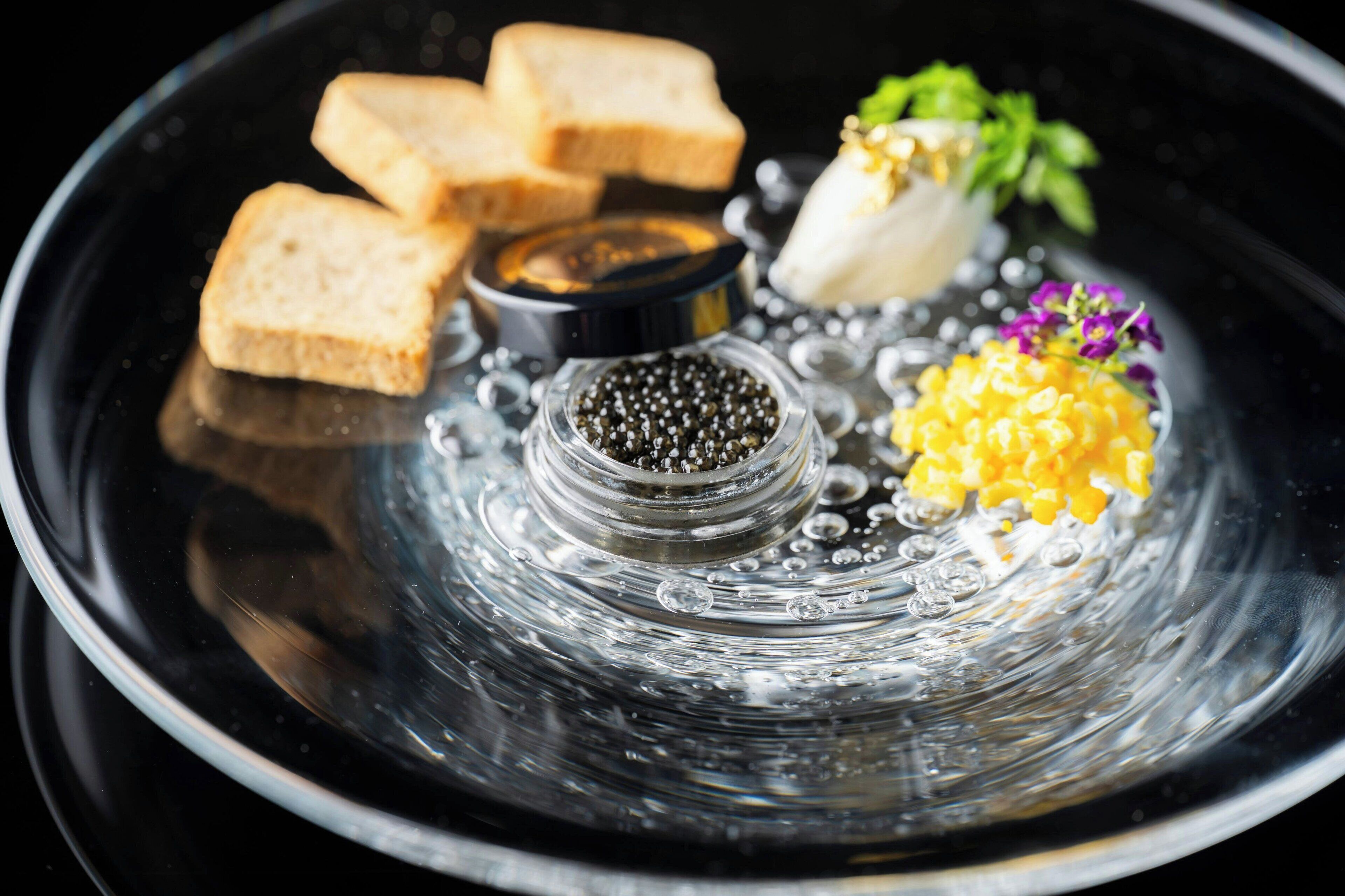 A clear glass bowl with caviar, lemon zest, and a small bouquet of flowers on a reflective surface.