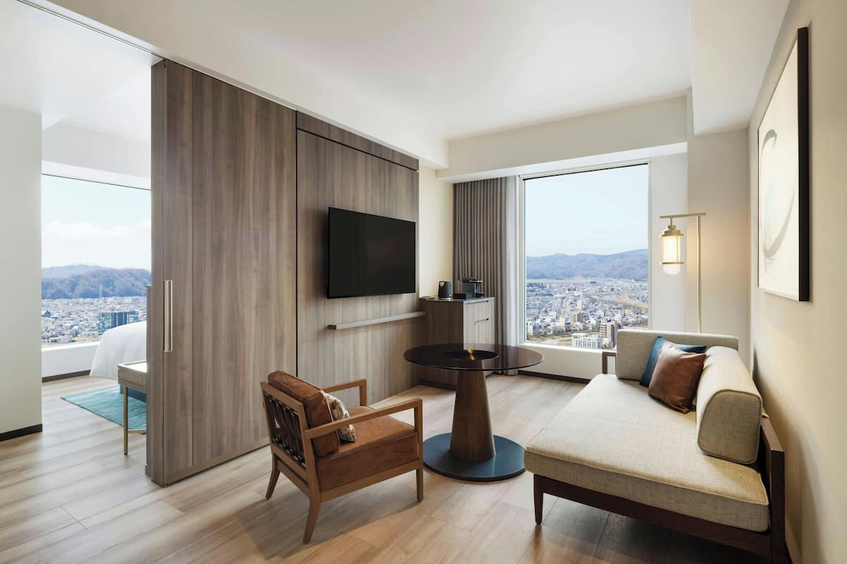 Ett modernt hotellrum med en soffa, en fåtölj, ett litet bord, en platt-TV och utsikt över staden.