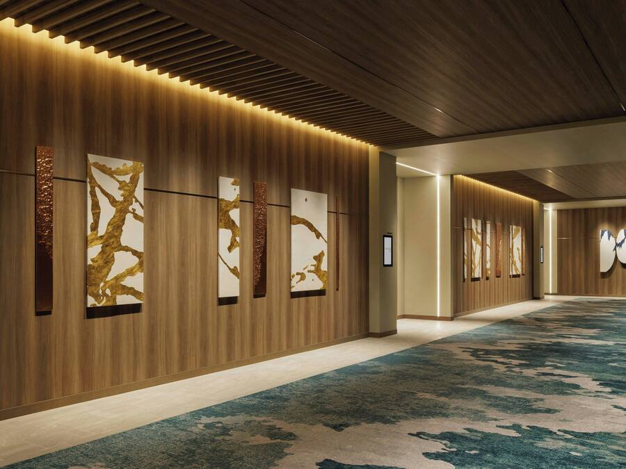 A modern hotel corridor with wooden panel walls, large abstract art pieces, and a patterned carpet.