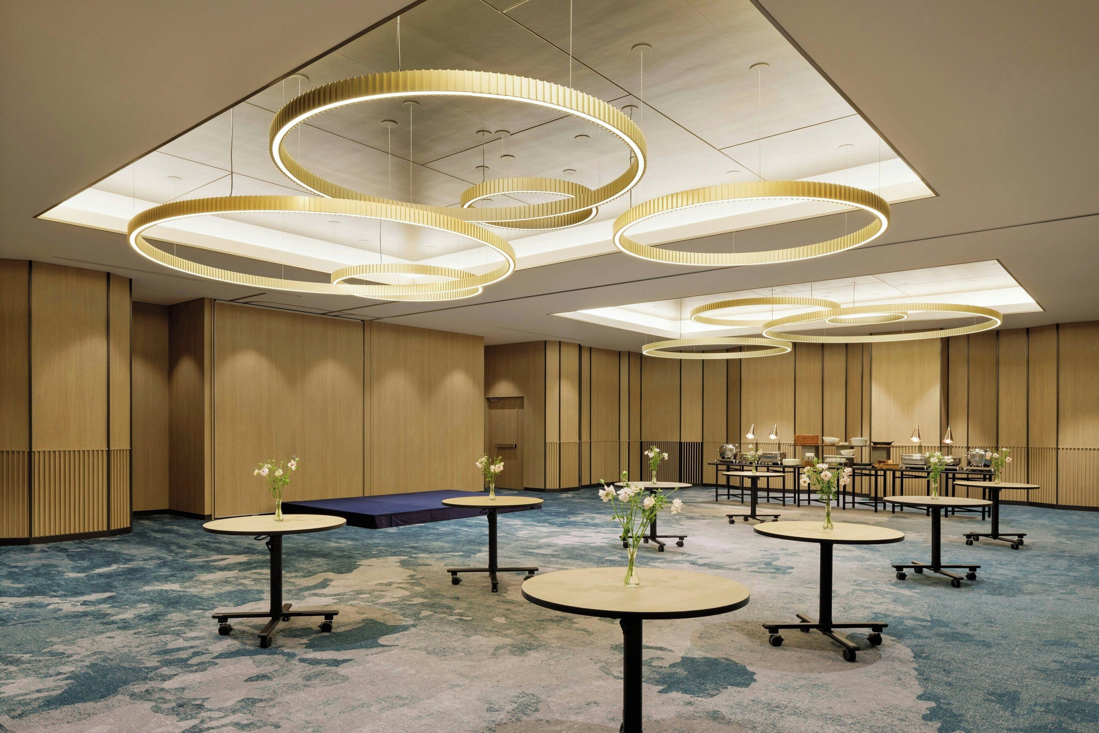 A modern hotel room with circular ceiling lights, round tables with flower vases, and a blue carpet.