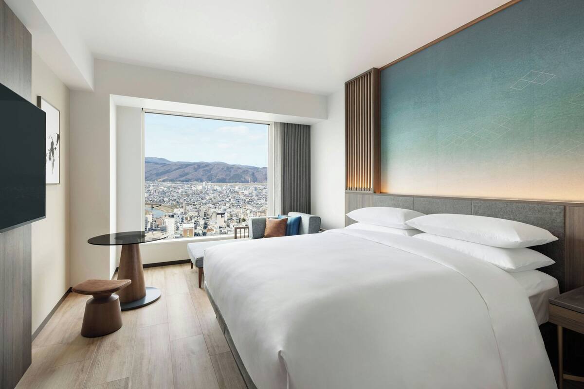 Ett modernt hotellrum med en stor säng, ett litet bord och utsikt över stadsbilden genom ett stort fönster.