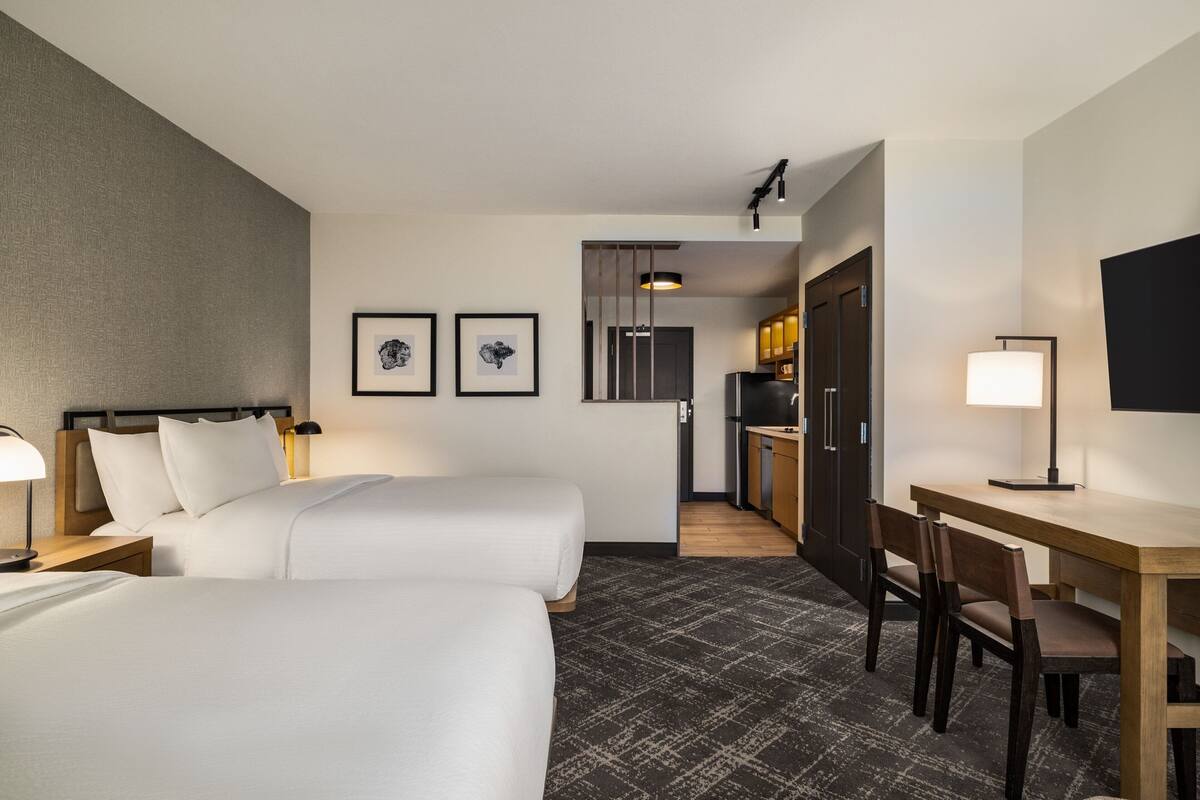 Habitación de hotel con dos camas, un escritorio, una silla, una lámpara y un televisor. Se ve un área de cocina a través de una puerta.
